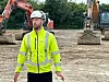 Mand i gul refleksvest og med hjelm på hovedet foran et par gravemaskiner.