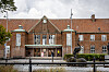 Billedet viser Nakskov Station set fra hovedindgangen og med statuen i forgrunden. Bygningen er i røde mursten.