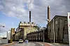 Billedet viser Sukkerfabrikken i Nakskov set fra gadeplan. I forgrunden er der en modkørende bil, imens der i baggrunden er fabrik og skorstene.
