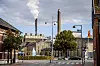 Billedet viser Sukkerfabrikken i baggrunden med træer og bygninger i forgrunden. Der er to skorstene i den øverste del og trafik fra biler på gaden i den nederste del af billedet.