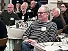Mennesker sidder ved borde med navneskilte og glas under et møde i et konferencelokale.