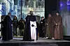 Skuespillere i munkekutter står på en oplyst teaterscene med kirkeligt bagtæppe.