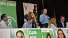 Panel af politikere fra flere partier taler ved et vælgermøde med kampagneplakater foran sig.