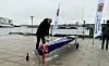 Person døber en blå robåd på kajen ved Nykøbing F. Roklub med vand og flag.