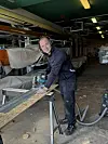 Håndværker saver en træplade på et bord i en bådehal med robåde i baggrunden.
