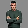 Portræt af mand i mørkegrøn trøje med korslagte arme foran grå baggrund.