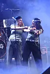 To performere danser synkront på en røgfyldt koncertscene med mikrofoner og trommesæt bag dem.