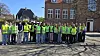 En gruppe personer i gule sikkerhedsveste står foran en murstensbygning på en solrig dag.