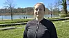 Kvinde står foran en sø i en park med træer og mennesker i baggrunden.