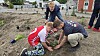 To børn planter små planter i jord ved en plads under anlæg, mens en voksen ser til.