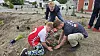 To børn planter små planter i jord ved en plads under anlæg, mens en voksen ser til.
