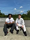 To mænd sidder på en betonkant foran en græsmark og hæk i solskin.