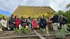 Mennesker står på en træsti foran et stråtækt skur dækket af gule blomster.