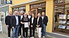 Otte personer står foran en butik med gul facade, og to af dem holder et papir ved døren.