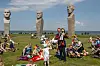 Mennesker sidder og står på en græsmark blandt høje stenfigurer under en blå himmel.