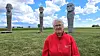 En ældre person i rød jakke står foran tre høje skulpturer på en græsmark under en blå himmel.
