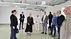 En gruppe mennesker står i et lyst industrilokale med højt loft og store vinduer.