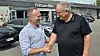 To mænd giver hånd foran en bilforretning med parkerede biler og skiltet Frihmann Biler.