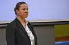 Kvinde i mørkt jakkesæt står foran en projektionsskærm i et indendørs lokale.
