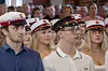 Fire unge mennesker sidder i et publikum og kigger fremad med kasketter i røde, hvide og sorte farver.