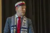 Mand i kasket og halstørklæde taler foran mørk baggrund.