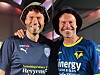 To mænd i fodboldtrøjer står sammen og smiler indendørs.