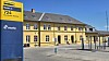 Gul stationbygning med blå Movia-stander og perronområde under blå himmel.
