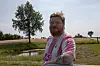 Mand i lyserød trøje tager selfie ved vandpyt i åbent land med træ og marker.
