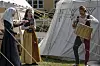 Tre personer i historiske kostumer står ved et telt og arbejder med reb og kurve i en udendørs lejr.