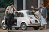 Tre personer ved en sølvfarvet Fiat 500 foran en bygade med en åben bildør.