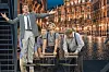Mandlig performer med udstrakte arme foran tre andre på en scene med en oplyst bykulisse.