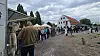 Folk står i kø på en grusplads foran en hvid historisk bygning.
