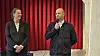 To mænd står på en scene; den ene taler i mikrofon foran et rødt scenetæppe.