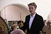 Mand i mørk jakke taler foran et publikum i en lys sal med et stort strengeinstrument.