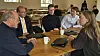 Gruppe voksne sidder samlet ved et cafébord med kaffe og en bærbar computer.
