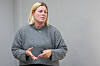 Blond kvinde i grå striktrøje gestikulerer foran en grå væg.