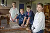 Fire unge står omkring et arbejdsbord i et værksted med værktøj og træmaterialer.