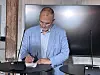 Mand i lyseblå blazer skriver ved et bord foran en stor skærm.