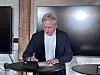 Gråhåret mand skriver ved et rundt bord foran to store skærme.