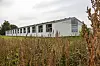 Lav moderne bygning ses bag høje, visne planter i et åbent landskab.