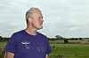 Gråhåret mand i lilla T-shirt foran grønne marker under en overskyet himmel.