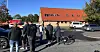 Gruppe mennesker med cykler på en parkeringsplads foran en bygning med rødt tag.