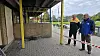 To personer står bag rød-hvid afspærring ved en murstensbygning og et udendørs anlæg.