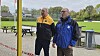 To mænd i sportsjakker står ved kanten af en græsbane.